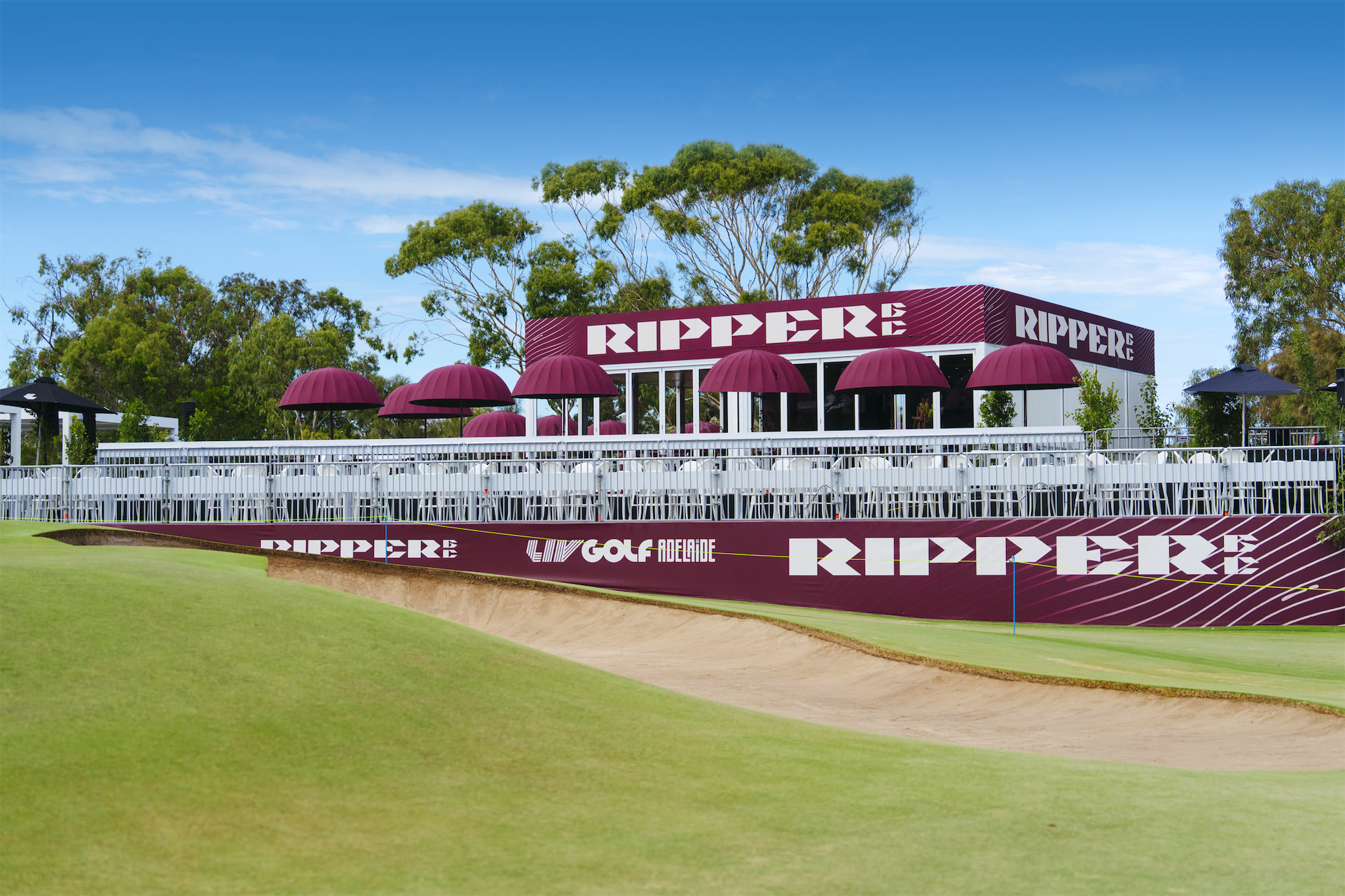 a branded modular event structure with a deck area filled with purple umbrellas, tables and chairs. Adjacent to a golf bunker.