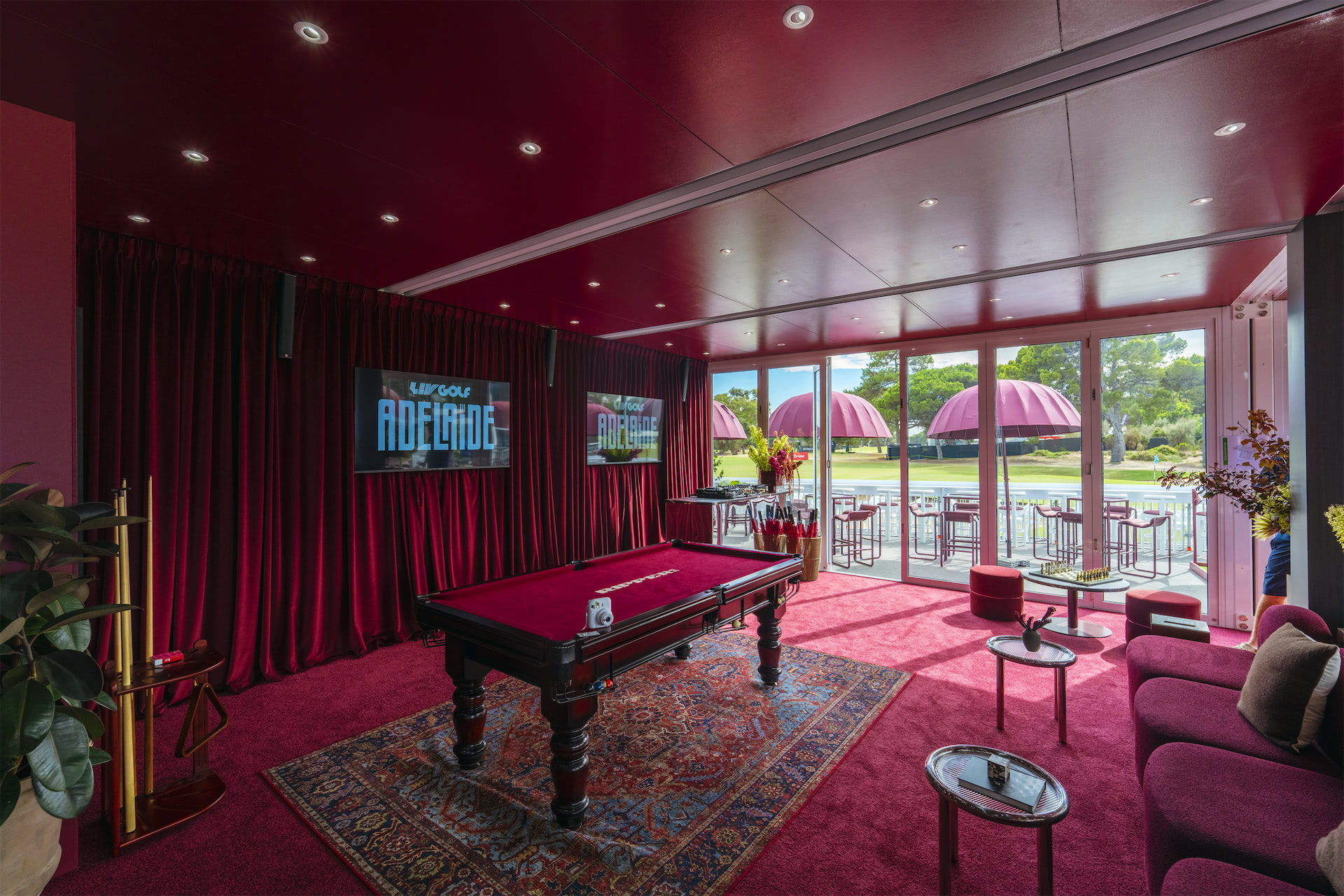 a pool table in a room with mauve curtains and carpets and big screen TV's on the wall. A view to the golf course outside.