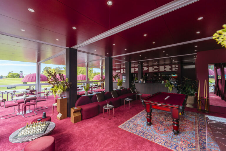 a pool table in a room with red carpet. Lounge seats and a bar along the back wall.