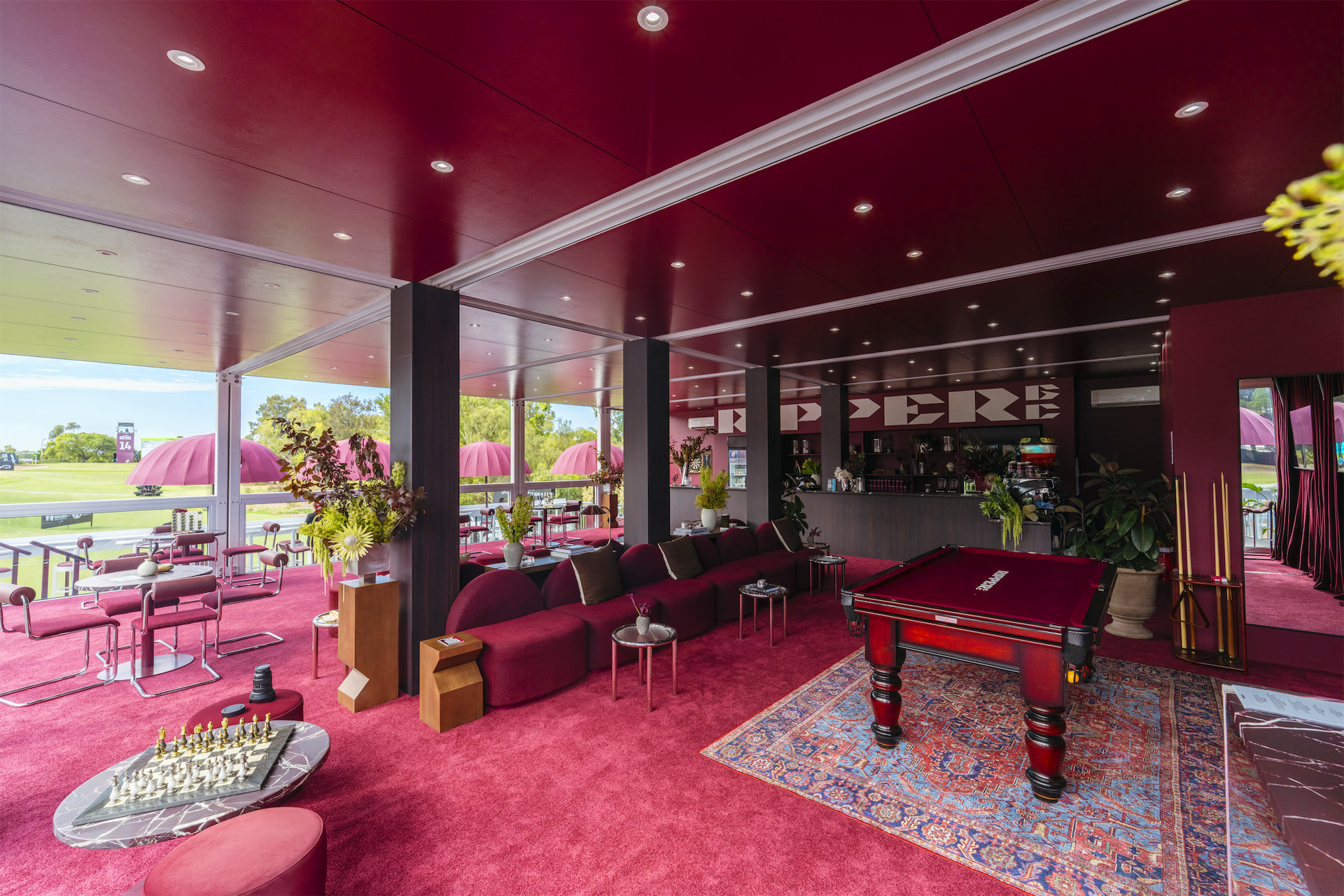 a pool table in a room with red carpet. Lounge seats and a bar along the back wall.